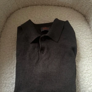 Mörkbrun merinoull pikétröja Morris - Snygg mörkbrun pikétröja från Morris i 100% merinoull. Klassisk krage och knäppning med två knappar framtill. Perfekt för dig som gillar stilrena och tidlösa plagg med en mjuk och lyxig känsla.