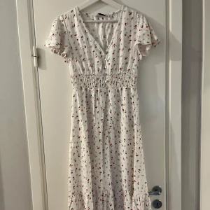 Vit blommig långklänning med v-ringning - Superfin vit långklänning från vero Moda med små röda och gröna blommor. Klänningen har v-ringning, knappar framtill, smock i midjan och korta ärmar med volang. Perfekt för sommarens alla tillfällen!