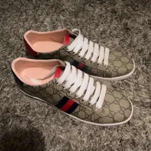 sneakers med monogram och röd/blå detalj - Snygga sneakers med klassiskt GG-monogram i beige och brunt, vita snören och vit sula. Sidan har den ikoniska röd-blå randen och insidan är ljusrosa. Perfekta för dig som vill sticka ut med lyxig streetstyle.