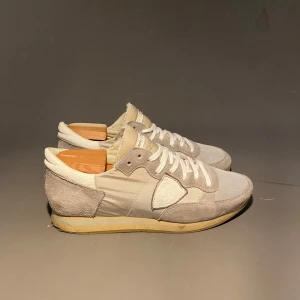 Philippe Model vita sneakers i mocka - Snygga vita sneakers från Philippe Model med detaljer i mocka och textil. Skorna har klassisk snörning, rund tå och en beige sula som ger en retrovibe. Perfekta för dig som gillar stilrena och neutrala sneakers med premiumkänsla.