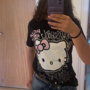 Hello Kitty svart t-shirt FB Sister - Svart t-shirt från FB Sister med stor Hello Kitty-tryck framtill och graffitiinspirerad text. Söt rosa rosett på Hello Kitty och coola vita och grå detaljer. Klassisk passform och kortärmad modell, perfekt för dig som gillar kawaii och streetstyle. Inte mycket använd kommer från new yorker🥰