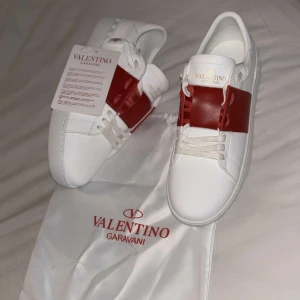 Valentino Garavani Open - sneakers, vinröd/vit - Helt nya, fräscha Valentino Garavani Open sneakers i vitt skinn med en bred vinröd läderdetalj över mitten. Klassisk rund tå, platt sula och coola nitar baktill på sulan. Skorna har vita snören och diskret logga på plösen. Perfekta för dig som gillar lyxig streetstyle.