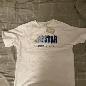 Vit Trapstar t-shirt med broderad logga - Storlek S men passa M. Vit t-shirt från Trapstar med broderad logga i blå och ljus grå på bröstet. Klassisk rund hals och korta ärmar. Helt ny, aldrig använd. Snygg streetwear-vibe med tydlig text och cool färgövergång i brodyren. Perfekt för dig som gillar statement-plagg.