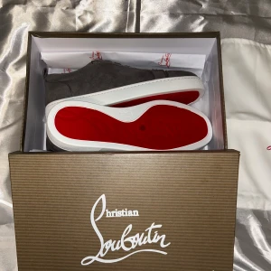 Grå mocka sneakers från Louboutin - Snygga grå sneakers i mocka från Christian Louboutin med klassisk röd sula och vita kanter. Skorna har rund tå, snörning och en stilren design som sticker ut med den ikoniska röda undersidan. Perfekta för dig som vill ha något exklusivt och trendigt.