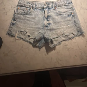 Ljusblå jeansshorts från Gina Tricot - Snygga ljusblå jeansshorts från Gina Tricot med fransig och sliten look. Klassisk femficksmodell och råa kanter nertill. Perfekta för sommardagar och festivaler. Styla med en crop top eller hoodie för en chill vibe.