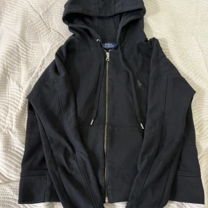 Svart kofta - Svart hoodie med dragkedja framtill och klassisk huva med snörning. Tröjan har långa ärmar, två fickor och diskret broderad logga på bröstet. Tillverkad i mjuk bomull för en skön och avslappnad stil. Storlek s funkar till xs också. 