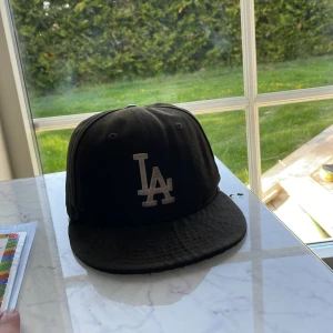 Svart LA-keps från New Era 59FIFTY - Snygg svart keps från New Era 59FIFTY med LA-logga broderad framtill i vitt. Klassisk flat brim och strukturerad passform. Perfekt för dig som gillar streetstyle och sportiga vibes.