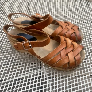Bruna flätade trä sandaler i skinn - Snygga sandaler i brunt skinn med flätade remmar och justerbart spänne vid ankeln. Träliknande sula med liten klack och öppen tå ger en retrovibe. Perfekta för varma dagar och ger en cool touch till din sommarstil. Använda, se bilderna, pris kan diskuteras!