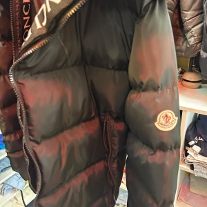 Svart Moncler dunjacka med logga - Snygg svart dunjacka från Moncler med klassisk logga på ärmen och stora, vadderade sektioner. Jackan har dragkedja framtill och coola Moncler-detaljer längs insidan. Perfekt för dig som vill ha en varm och trendig vinterjacka.