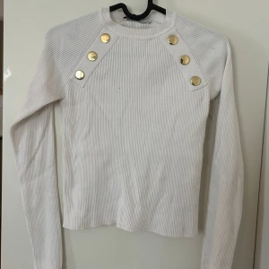 Vit ribbad tröja med guldknappar Zara - Supersnygg vit ribbad tröja från Zara med lång ärm och coola guldknappar på axlarna. Tröjan har en tight passform och är perfekt för dig som gillar stilrena detaljer. Materialet är mjukt och stretchigt, vilket gör den riktigt skön att bära.