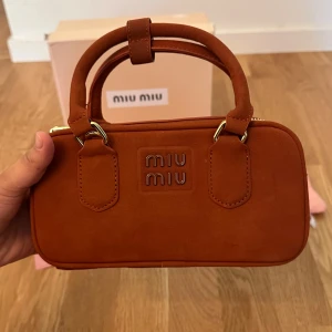 Brun handväska i mocka från Miu Miu - Supersnygg brun handväska från Miu Miu i mjuk mocka med guldiga metalldetaljer och dubbla handtag. Väskan har stilren, rektangulär form, präglad logga framtill och dragkedja upptill. Perfekt storlek för det viktigaste och ger en lyxig vibe. Helt ny 