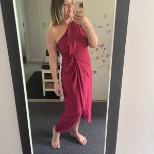 Röd oneshoulder långklänning - Säljer en snygg röd långklänning med oneshoulder och cool knutdetalj framtill. Klänningen har en draperad look och är perfekt för dig som vill sticka ut. Materialet känns mjukt och lätt, och modellen är figurnära men ändå bekväm.Tyvärr kan jag inte knäppa den,den är förliten för mig (syns på bilderna).Helt ny