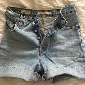 Levi's ljusblå jeansshorts hög midja - Säljer ett par klassiska ljusblå jeansshorts från Levi's med hög midja och raka ben. Shortsen har råa, uppvikta kanter och femficksmodell med knappar framtill. Perfekta för sommaren och ger en avslappnad vibe.