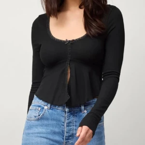 Svart croppad topp med rosett - Snygg svart croppad topp med lång ärm och liten rosett framtill. Toppen har v-ringning med spetskant och en lätt utsvängd nederdel som ger en cool siluett. Perfekt att styla med jeans eller kjol för en trendig look.