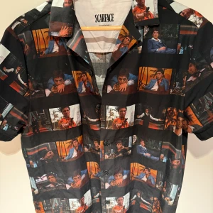 Scarface kortärmad skjorta med filmtryck - Unik svart skjorta med korta ärmar och knappar framtill, täckt av färgstarka tryck med ikoniska scener från filmen Scarface. Skjortan har klassisk krage och är perfekt för dig som vill sticka ut med en filmisk vibe.