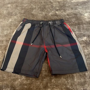 Burberry badshorts - Snygga badshorts med den klassiska rutigt Burberry mönster i svart, grått, beige och rött. Shortsen har elastisk midja med snörning och en bakficka. Perfekta för sommaren och strandhäng. Priset är ej hugget i sten vid snabb affär!