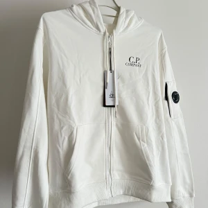 Vit hoodiejacka från C.P. Company - Vit hoodiejacka från C.P. Company med dragkedja framtill, två fickor och ikonisk linsdetalj på ärmen. Passar även för dem som har Storlek S men i rätt kroppsform 