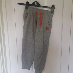 Grå mjukisbyxor Lupilu med badge - Grå mjukisbyxor från Lupilu med orange snörning i midjan och en cool badge med dinosaurietema på ena benet. Byxorna har ribbade muddar vid bensluten och är gjorda i mjukt bomullsmaterial. Perfekta för chill dagar eller lek.