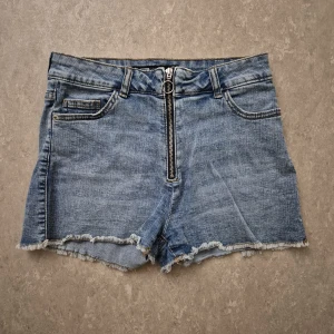 Jeansshorts - Snygga ljusblå jeansshorts från FB Sister med hög midja och råa, fransiga kanter. Klassisk femficksmodell med dragkedja framtill och bälteshällor. Perfekta för varma dagar och en avslappnad look.