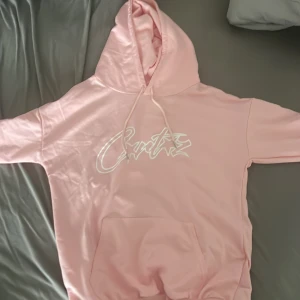 Rosa hoodie Xs - Säljer en ljusrosa hoodie  i storlek XS. Tröjan har vit logga framtill, stor känguruficka och huva med snörning. Perfekt för dig som gillar en chill och snygg streetstil.