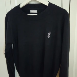 Svart tröja, Yves Saint Laurent, XS - Svart stickad tröja från Yves Saint Laurent med klassisk rund halsringning och broderad YSL-logga på bröstet. Tröjan har långa ärmar och ribbade muddar. Storlek: XS 