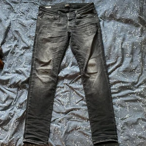 Slim svarta jeans Jack & Jones - Svarta slim fit jeans från Jack & Jones i modellen Slim/Glenn. Jeansen har egengjorda slitna detaljer på låren och knäna. Snyggt tvättade med en mörkgrå ton och stretchig bomullsblandning för extra komfort.