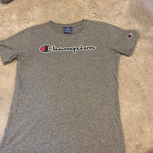 Grå Champion t-shirt med logga - Snygg gråmelerad t-shirt från Champion med stor vit och röd logga över bröstet och liten logga på ärmen. Klassisk rund hals och korta ärmar. Perfekt för en chill och sportig stil.