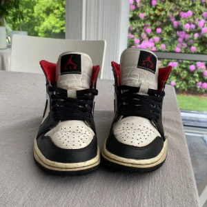 Nike Air Jordan 1 svart/vit/röd 36.5 - Nike Air Jordan 1 sneakers i svart, vitt och rött skinn. Klassisk high-top siluett med svart swoosh, röda detaljer vid ankeln och svart snörning. Perforerad tåbox och ikonisk Air Jordan-logga på sidan. Perfekta för dig som gillar streetstyle och retro vibes.