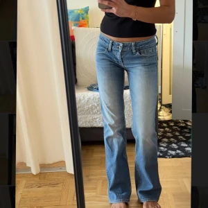 Blå bootcut jeans  - Säljer ett par ljusblå bootcut jeans med klassisk femficksdesign och snygga slitningar. Jeansen har låg midja, raka ben med lätt utsvängning och detaljerad bakficka med A-logga på läderpatch. Perfekta för en avslappnad och trendig look.
