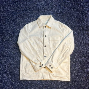 Vit overshirt från Arket i bomull - Snygg vit overshirt från Arket i bomullstwill. Jackan har klassisk krage, knäppning framtill med svarta knappar och tre fickor – två stora nedtill och en bröstficka. Perfekt lager-på-lager-plagg med clean look och raka linjer. Finns en fläck som inte är så synlig på bild nr.5. Hör av er vid minsta fundering🙌
