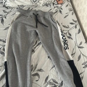 Grå mjukisbyxor Jack & Jones - Grå mjukisbyxor från Jack & Jones med svarta och vita paneler längs sidorna och logga på ena benet. Byxorna har resår i midjan med snörning och är tillverkade i mjukt bomullsmaterial. Perfekta för chill eller träning.
