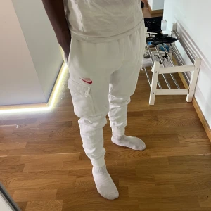 Vita cargo mjukisbyxor från Nike - Säljer ett par vita mjukisbyxor från Nike med cargo-fickor på sidorna och röd Nike-logga. Byxorna har resår i midjan och vid bensluten för en skön passform. Perfekta för chill eller streetwear-stil.