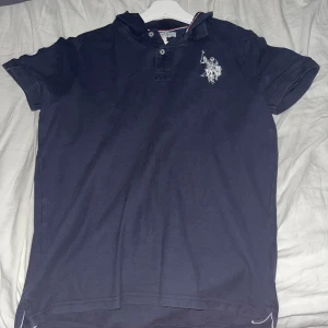 Mörkblå pikétröja från U.S. Polo Assn. - Säljer en klassisk mörkblå pikétröja från U.S. Polo Assn. med vit broderad logga på bröstet. Tröjan har korta ärmar, krage med knappar och snygga detaljer i kragen med röd och vit rand. Perfekt för en stilren och sportig look, prefekt nu till sommaren.