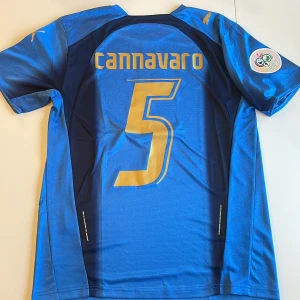 Italien Cannavaro VM 2006 tröja Puma - Italiens blåa fotbollströja replika från VM 2006 med Cannavaro och nummer 5 i guld på ryggen. Puma-logga på bröst och ärm, officiellt VM-märke på ärmen och Italiens landslagsemblem framtill. Tillverkad i lätt och ventilerande material, perfekt för fotbollsfans. Endast handtvätt!