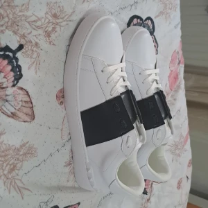 Valentino Garavani Open sneakers vit/svart - Snygga Valentino Garavani Open sneakers i vitt skinn med bred svart läderdetalj över vristen. Klassisk rund tå, vita snören och diskret guldlogga på plösen. Perfekt för dig som gillar stilrena och lyxiga sneakers med en modern twist.