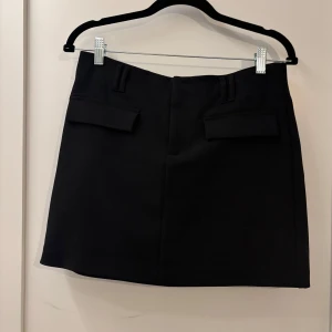 Svart kortkjol med fickdetaljer - Stilren svart kortkjol med raka linjer och två fickdetaljer framtill. 