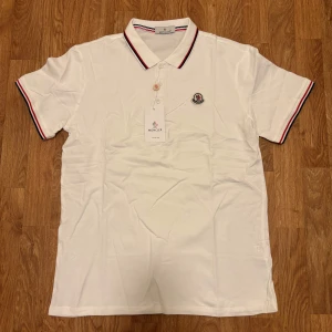 Vit Moncler pikétröja med ränder - Snygg vit pikétröja från Moncler med klassisk krage och knäppning framtill. Tröjan har Moncler-logga på bröstet och röda samt svarta ränder på krage och ärmslut. Tillverkad i bomull för en skön och stilren känsla.