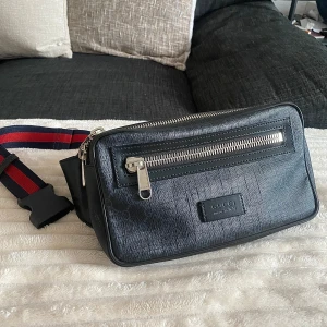 Svart Gucci magväska med GG-mönster - Säljer en svart magväska från Gucci med klassiskt GG-mönster och silverfärgade dragkedjor. Väskan har justerbart band i rött och marinblått, samt präglad logotyp på framsidan. Tillverkad i skinn med struktur och detaljer i textil. Perfekt för att bära det viktigaste med stil.