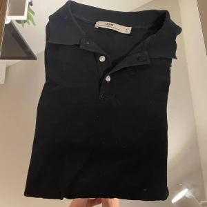 Svart pikétröja från HOPE - Klassisk svart pikétröja från HOPE med krage och två knappar framtill. Tröjan har korta ärmar och är tillverkad i mjuk bomull, perfekt för en clean och stilren look. Passar dig som gillar minimalistisk stil och vill ha något enkelt men snyggt.