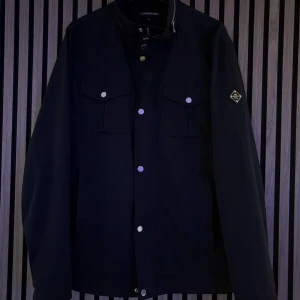 J LINDEBERG BAILEY JACKA  - En J Lindeberg jacka i klassiskt M65 utförande med gott om fickor invändigt som utvändigt.   Storlek L  Svart   Mycket bra skick. Inga defekter. Använt ett fåtal gånger.  Vattentät.  PM för mer bilder eller frågor.