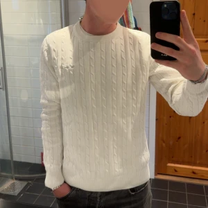 Vit kabelstickad tröja - Säljer en stilren vit kabelstickad tröja med rund hals och långa ärmar. Tröjan har klassiskt mönster och ribbade muddar vid ärmslut och nederkant. Perfekt för dig som gillar en clean och tidlös look.
