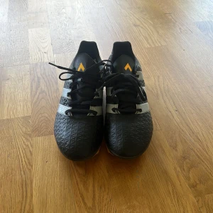 Adidas 16.4 svarta fotbollsskor - Svarta Adidas 16.4 fotbollsskor med klassiska tre silverfärgade ränder på sidorna och orange detaljer. Skorna har snörning och mönstrad ovandel i syntetmaterial för extra grepp. Perfekta för dig som vill sticka ut på planen. Har använt dom 3 gånger. Skriv på för frågor☺️