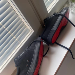 Svarta sneakers med röd sula - Snygga svarta sneakers från Christian Louboutin med ikonisk röd sula och svarta snören. Ovandelen är i mocka och insidan har en mjuk, ljusbrun läderdetalj. Skorna har rund tå och klassisk sneakerlook med metallöljetter.