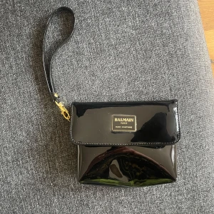 Svart kuvertväska från Balmain - Snygg svart kuvertväska från Balmain med glansig finish och guldfärgad logga framtill. Väskan har avtagbar handledsrem och stängs med magnetknapp. Insidan är fodrad i neon-gult tyg som ger en cool kontrast. Perfekt för att bära det viktigaste.