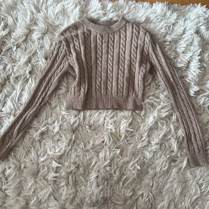 Beige croppad kabelstickad tröja - Superfin croppad kabelstickad tröja i beige med rund hals och långa ärmar. Tröjan har bred ribbad mudd vid ärmslut och nederkant, perfekt för lager-på-lager och snygg till både jeans och kjol.