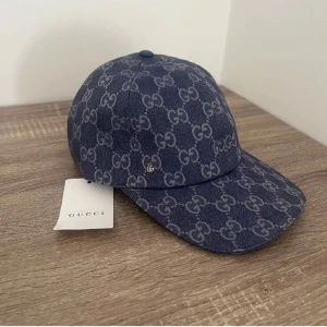 Blå keps från Gucci med GG-mönster - Snygg blå keps från Gucci med klassiskt GG-monogram i grått över hela kepsen. Justerbar rem baktill i blått skinn och guldfärgade detaljer. Tillverkad i bomullstyg med exklusiv känsla och perfekt passform. En riktig statement-accessoar för dig som gillar lyxiga märken.