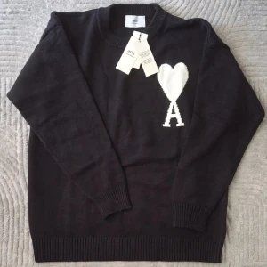 Svart stickad tröja från A - Svart stickad tröja från  Alexandre Mattiussi med vit hjärt- och A-logga på bröstet. Tröjan har rund hals, ribbade muddar och lång ärm. Perfekt för dig som gillar stilrena detaljer och vill ha en snygg statement piece i garderoben.