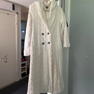 Vit lång teddyjacka med huva - Supermysig lång teddyjacka i vit färg med huva och dubbelknäppning framtill. Jackan har ett stickat kabelmönster i tyget och stora knappar som ger en cool look. Perfekt för kalla dagar när du vill vara både varm och stylish.Helt ny