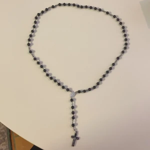 Svart pärlhalsband med kors - Snyggt halsband med svarta pärlor och ett silverfärgat kors som hänger ned. Kedjan är tunn och pärlorna är jämnt fördelade runt hela halsbandet. Perfekt för dig som gillar stilrena och lite edgy accessoarer.