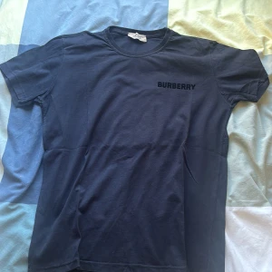 Mörkblå Burberry t-shirt med logga - Oanvänd Snygg mörkblå t-shirt från Burberry med klassisk logga tryckt på bröstet och ett stort, mönstrat Burberry B på ryggen. T-shirten har rund hals och korta ärmar, tillverkad i mjuk bomull. Perfekt för dig som gillar stilrena designer med en twist.